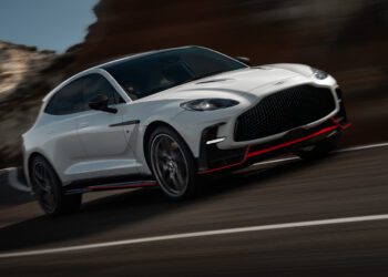 A white Aston Martin DBX S Super-SUV with black and red accents is driving at speed on a curved mountain road.