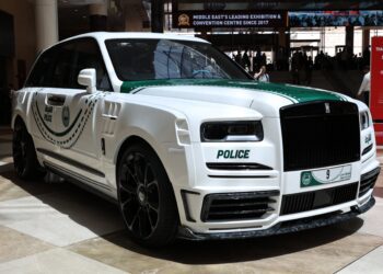 A white Rolls-Royce SUV with green police markings and emblems is displayed indoors, featuring a Dubai police license plate.
