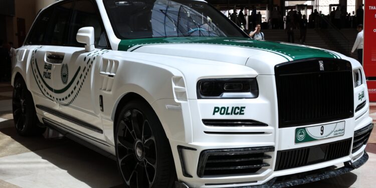 A white Rolls-Royce SUV with green police markings and emblems is displayed indoors, featuring a Dubai police license plate.