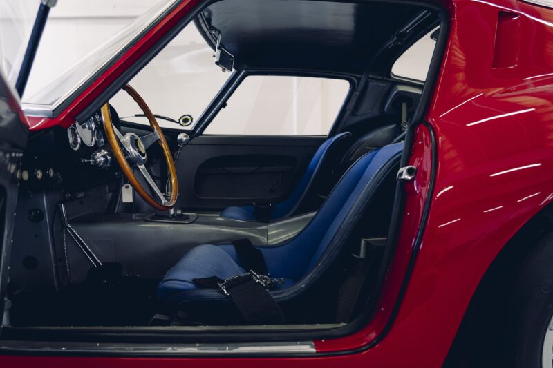 Interior view of a classic red Ferrari 330 LM/250 GTO sports car with blue bucket seats, a wooden steering wheel, and a manual gear shift—an icon you might see at the Cavallino Classic 2025.