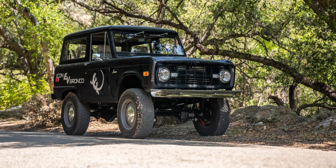 A vintage black off-road vehicle is parked on a forested road, surrounded by trees and dappled sunlight.