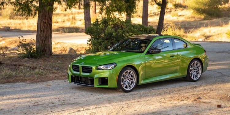 A bright green BMW coupe is parked on a dirt road near trees and dry grass in a sunlit outdoor setting.