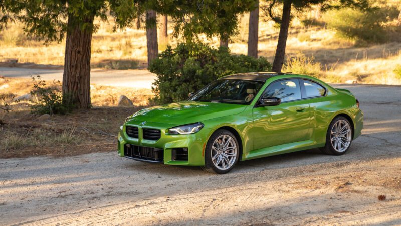 A bright green BMW coupe is parked on a dirt road near trees and dry grass in a sunlit outdoor setting.