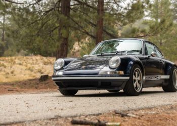 A classic dark blue Porsche 911 is parked on a forest road, surrounded by trees and natural scenery.