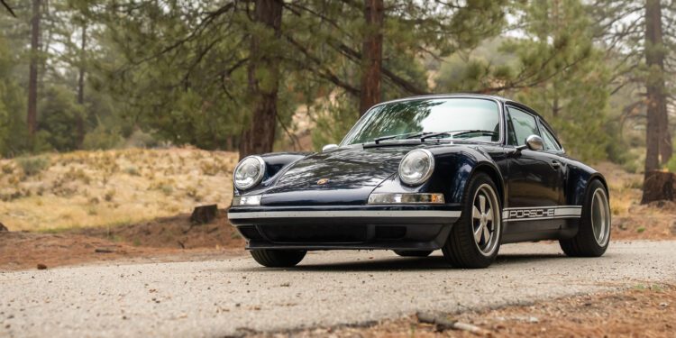 A classic dark blue Porsche 911 is parked on a forest road, surrounded by trees and natural scenery.