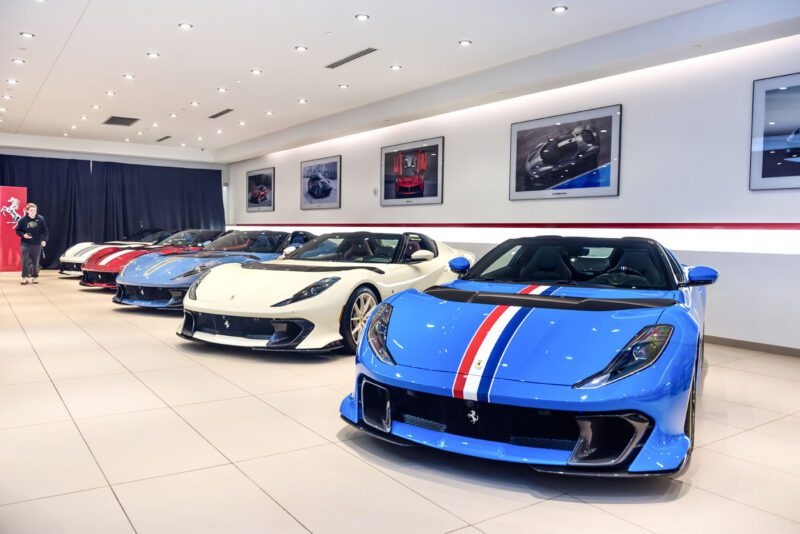 A stunning exhibition features a row of luxury sports cars, including striking blue, white, and red models like the Ferrari 812 Competizione, all elegantly displayed in the showroom amidst framed car photos on the wall.