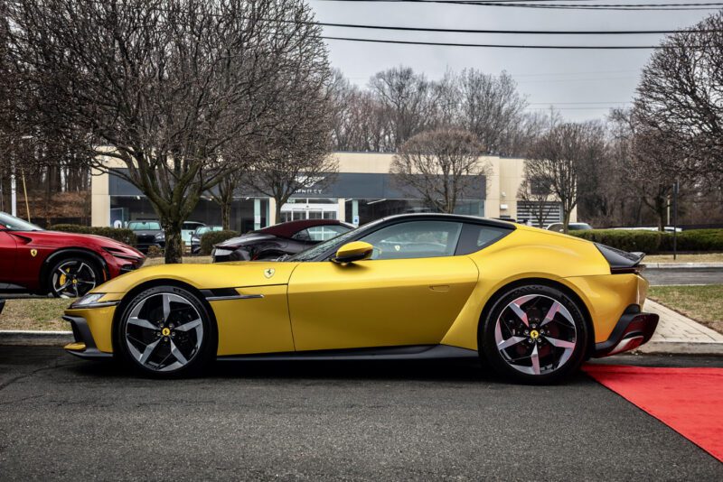 A sleek yellow Ferrari 812 Competizione graces a Central New Jersey street, parked elegantly beside a red carpet with bare trees standing silently in the background.