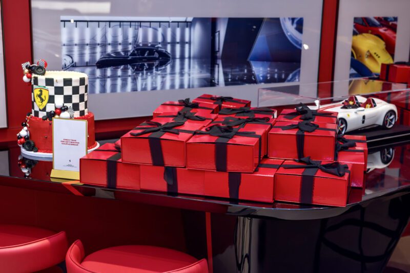 Red gift boxes with black ribbons stacked on a table next to a decorative cake featuring car elements and a small race car model bring the excitement of an exhibition to life, complemented by Ferrari 812 Competizione-themed wall art in the background.