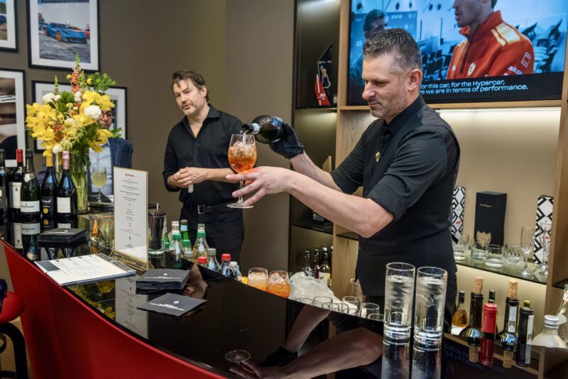 A bartender serves a drink at a bar adorned with flowers and assorted spirits, evoking the elegance of a Ferrari exhibition. Nearby, another man stands captivated by the scene, much like an enthusiast admiring the sleek lines of a Ferrari 812 Competizione.