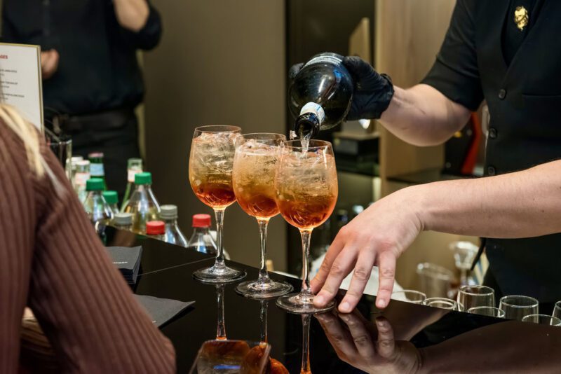 A bartender, like an artist at a Ferrari exhibition, pours sparkling wine into three glasses of orange-colored cocktails on a sleek bar counter.