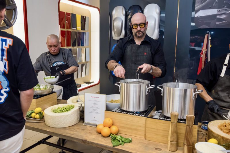 Two chefs at a modern kitchen station expertly prepare dishes with large pots and fresh ingredients, like lemons and greens, as if putting on a culinary exhibition worthy of admiration, while a guest overflows with excitement much like when first beholding the sleek Ferrari 812 Competizione.