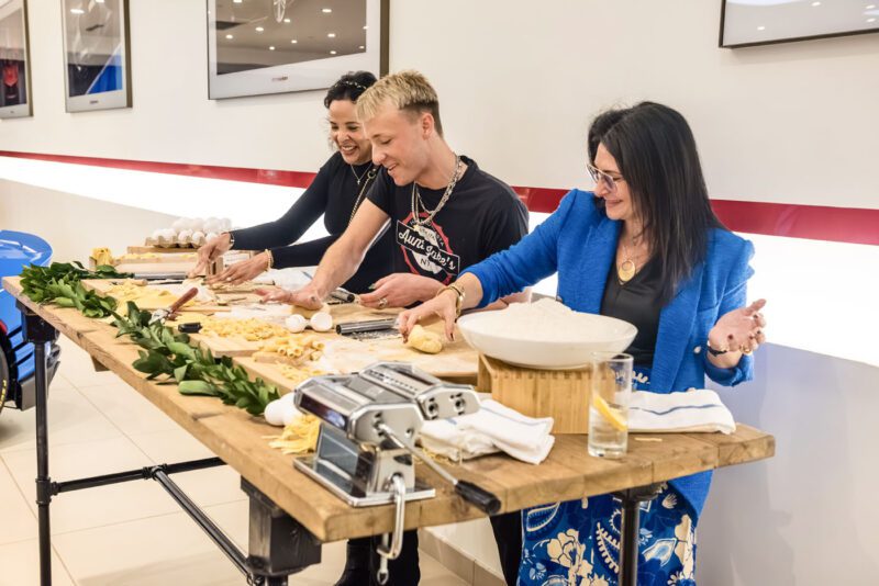 Three people making pasta at a wooden table, surrounded by pasta machines, eggs, and dough. Smiling and enjoying the activity, their laughter could rival an exhibition at Ferrari Central New Jersey.
