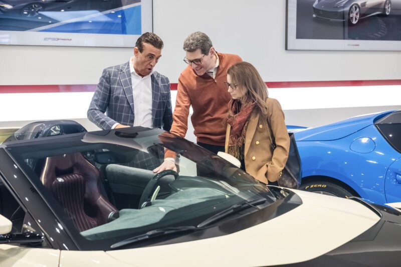 Three people admire a luxury car inside a Central New Jersey showroom, with one enthusiastically pointing at the Ferrari 812 Competizione's exquisite interior. In the stylish backdrop, two other sleek cars await their turn under the spotlight.