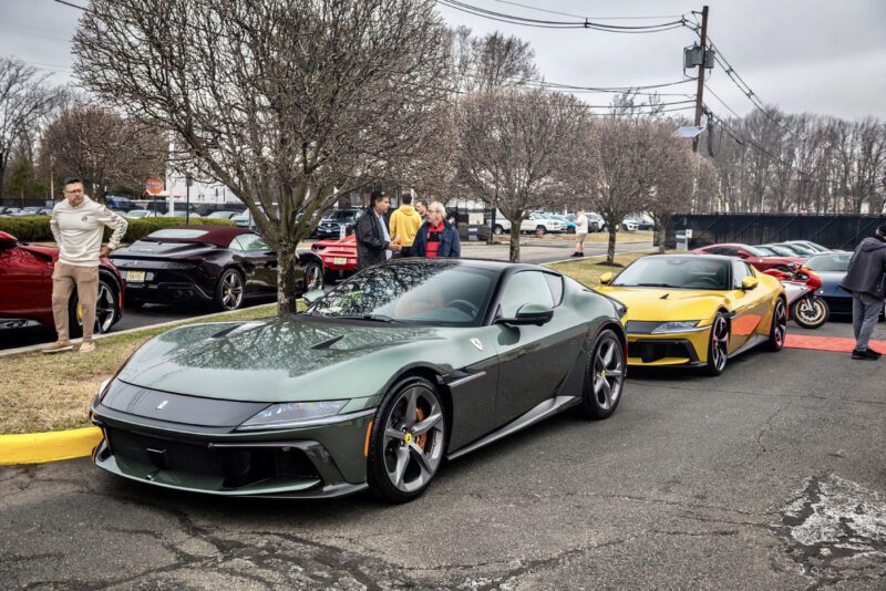 A sleek green sports car is parked on the street with a gleaming yellow Ferrari 812 Competizione behind it, creating an impromptu exhibition. Several people stand nearby under leafless trees and cloudy skies, admiring the stunning display.