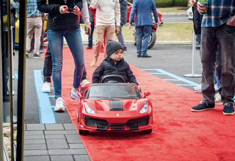 A child in a red toy car zips around a red carpet, capturing the attention of adults as if starring in an exhibition at Ferrari Central New Jersey.