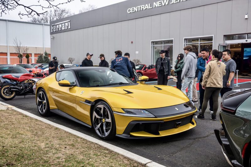 A yellow Ferrari 812 Competizione steals the spotlight at an exhibition, parked in front of a dealership and drawing a crowd despite the cloudy day. In the background, other cars and a motorcycle await their turn in the limelight.
