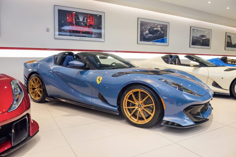 A Ferrari 812 Competizione with dazzling blue paint and striking yellow wheels stands out in a Central New Jersey showroom, framed by sleek car images on the walls.