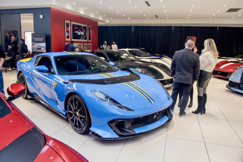 At Ferrari Central New Jersey, a striking blue Ferrari 812 Competizione with bold yellow stripes dazzles in the showroom exhibition, while enthusiasts admire the car amidst an array of stunning vehicles in the background.