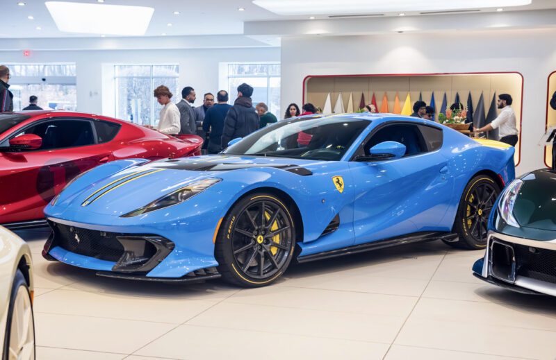 A stunning blue sports car with yellow accents, reminiscent of the Ferrari 812 Competizione, draws all eyes at the exhibition in Ferrari Central New Jersey's showroom, surrounded by an admiring crowd.