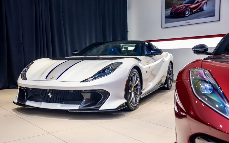A stunning white sports car with a dark racing stripe is showcased indoors beside a vibrant red Ferrari. In the backdrop, black curtains and a framed car photo add elegance to the exhibition, highlighting the impressive 812 Competizione's sleek design.