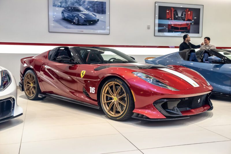 A striking red Ferrari 812 Competizione with white stripes and the number 16 on its side commands attention in the showroom. An eager crowd of car enthusiasts gathers in the background, captivated by this exquisite exhibition of automotive excellence.