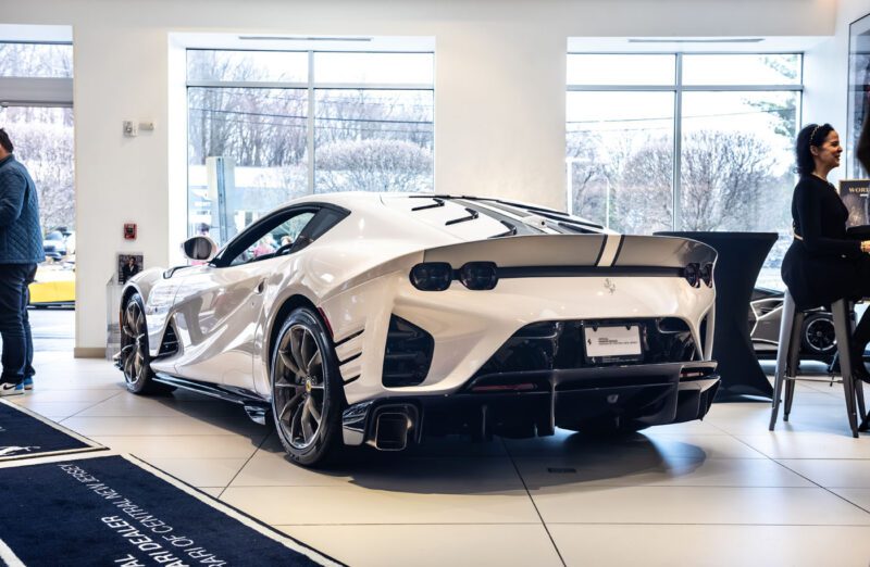 A stunning white Ferrari 812 Competizione with a sleek black spoiler is showcased in the exhibition at Ferrari Central New Jersey. Nearby, a woman stands confidently at the counter, ready to offer insights about this high-performance masterpiece.