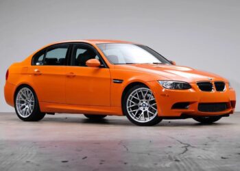 An orange BMW sedan with alloy wheels is parked indoors on a gray concrete surface against a plain light gray background, showcasing the collector-grade BMWs offered by Enthusiast Auto Group.