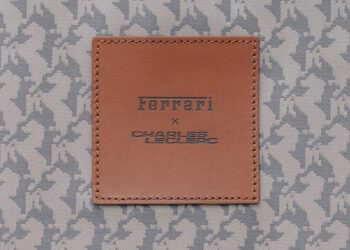 A brown leather patch with "Ferrari x Charles Leclerc" embossed, sewn onto gray fabric with a repeating horse pattern—a touch of luxury perfect for those who follow the latest lifestyle news.