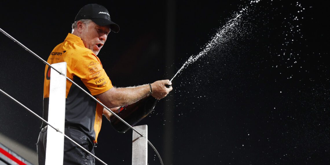 A jubilant person in team attire, perhaps celebrating a McLaren F1 victory, sprays champagne from a bottle on the platform.