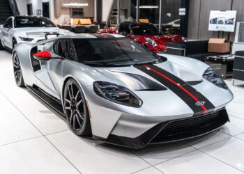 A silver Ford GT sports car with black and red stripes is displayed in a modern showroom with other luxury cars, showcasing one of the finest collectible Ford GTs for sale.