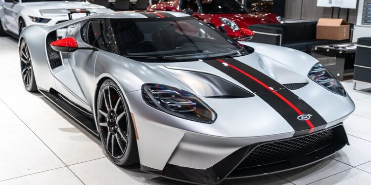 A silver Ford GT sports car with black and red stripes is displayed in a modern showroom with other luxury cars, showcasing one of the finest collectible Ford GTs for sale.
