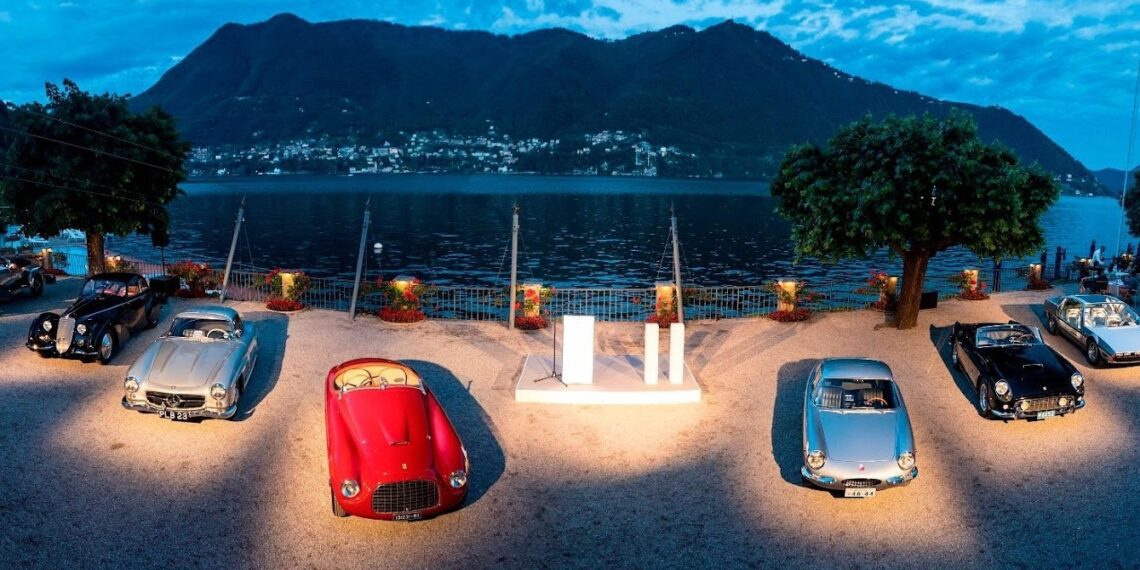 A group of classic cars displayed outdoors near a waterfront, with mountains and a small town in the background under a blue sky—an ideal setting for one of 2025’s top 10 car auctions.