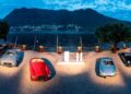 A group of classic cars displayed outdoors near a waterfront, with mountains and a small town in the background under a blue sky—an ideal setting for one of 2025’s top 10 car auctions.