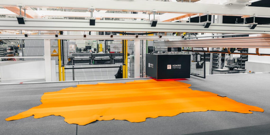 An industrial machine cuts a large piece of orange material in a factory setting, surrounded by metal structures and machinery.