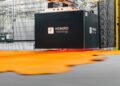 Orange material, resembling fine leather, is being precisely processed under a large black machine labeled "HOMIRO metrology," showcasing cutting-edge AI efficiency in a sophisticated factory setting.