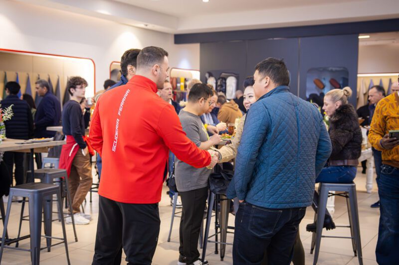 People socializing and mingling in a modern indoor setting, with some standing and others seated on bar stools, as the latest clothing collection is displayed in the background. Nearby, a Ferrari 812 Competizione steals glances, courtesy of Ferrari Central New Jersey's exclusive exhibition.