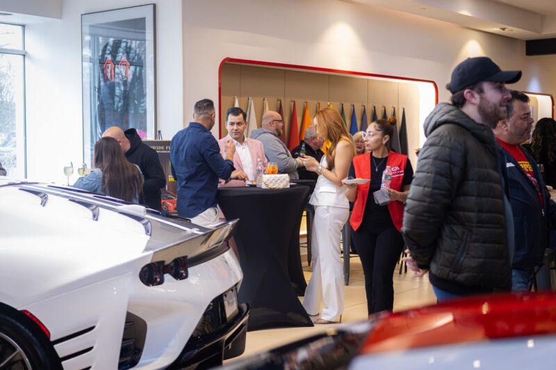 Guests are mingling at an exhibition held in a showroom, showcasing luxury cars like the Ferrari 812 Competizione. Some attendees, gathered around high tables with drinks and snacks, admire vibrant samples on the walls at Ferrari Central New Jersey.