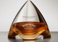 A uniquely shaped bottle of Bowmore single malt Scotch whisky with a silver cap, reflecting a modern design inspired by the sleek lines of the Aston Martin Valkyrie Hypercar on a neutral background.
