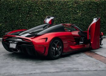 A red and black Koenigsegg sports car with both doors open is parked on a concrete driveway in front of a tall hedge—a stunning example of hypercars, possibly available for sale.