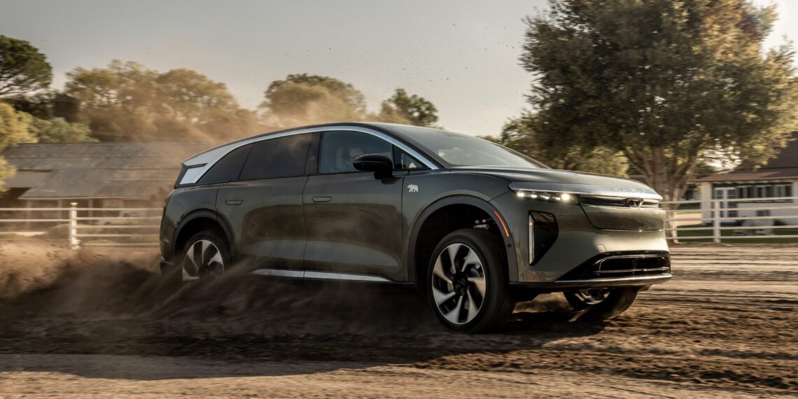 A dark SUV drives through dirt, raising dust, on a sunny day near wooden fences and trees.