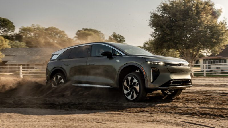 A dark SUV drives through dirt, raising dust, on a sunny day near wooden fences and trees.