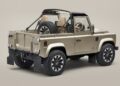 A beige two-door Classic Defender pickup with a soft top, open cargo bed, roll bar, and spare tire mounted behind the seats stands against a plain background.