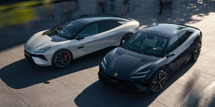 Two sleek sports cars, a white Lotus Emeya and a black Eletre, are parked next to each other on a paved surface, with the blurred motion of people bustling around them.