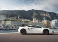 A white sports car is parked near a waterfront with mountains, modern buildings, and a cloudy sky in the background.