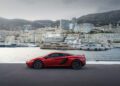 A red sports car, reminiscent of a McLaren auction highlight, is parked near a harbor with luxury yachts and high-rise buildings in the background, set against a mountainous landscape.