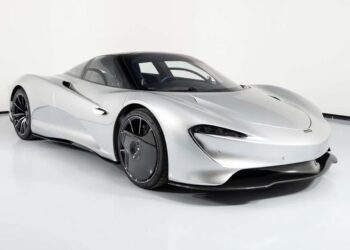 A silver McLaren Speedtail hypercar, inspired by the Electrified Longtail concept, is parked in a white studio, featuring a sleek, aerodynamic design with black accents and futuristic wheels.