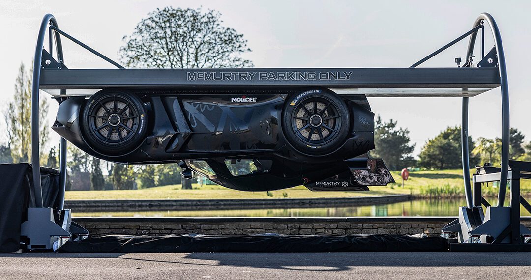 A McMurtry Spéirling is parked horizontally on a raised platform labeled "McMurtry Parking Only," creating the illusion of an almost gravity-defying stunt against a serene backdrop of trees and a pond.