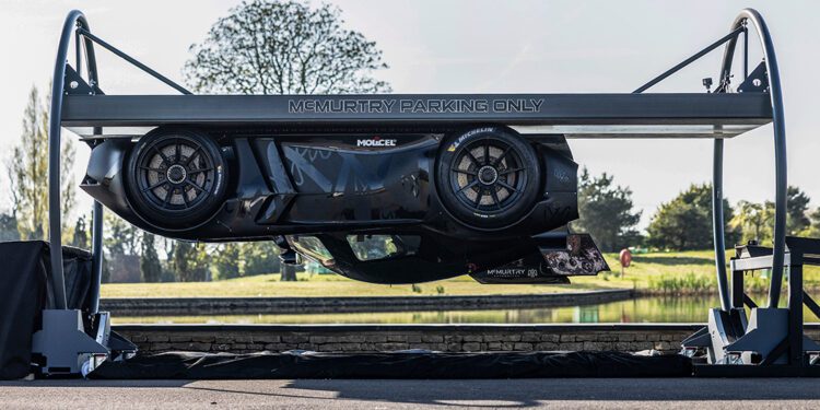 A McMurtry Spéirling is parked horizontally on a raised platform labeled "McMurtry Parking Only," creating the illusion of an almost gravity-defying stunt against a serene backdrop of trees and a pond.
