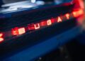 Close-up of the illuminated red Bugatti logo on a car's rear with a textured background, set against a hint of the iconic French Racing Blue.