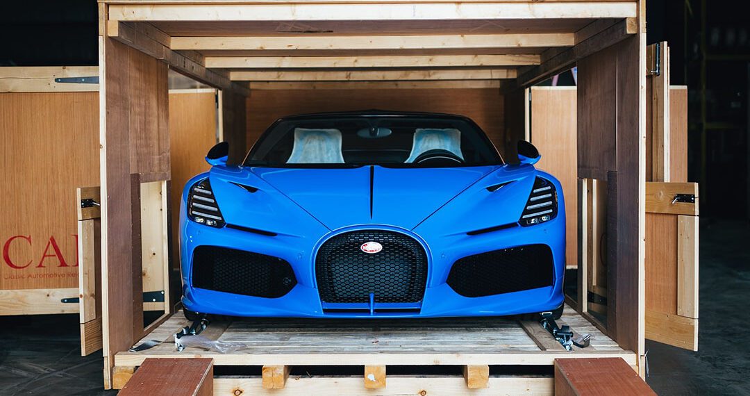 A Bugatti Mistral in striking French Racing Blue sits poised inside a wooden shipping crate, with its sleek front facing outward.